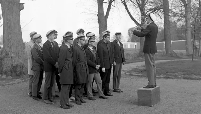 En man står på en fyrkantig låda framför en grupp människor på en grusgång. Alla har en studentmössa på huvudet. Träd i bakgrunden.