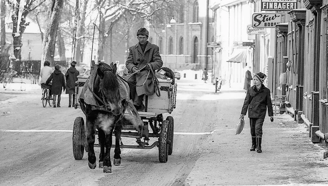 En väg där en person transporteras via häst och vagn. Ett barn går bredvid på gågatan. Butiker sträcker sig från förgrunden till bakgrunden. Några människor i bakgrunden.