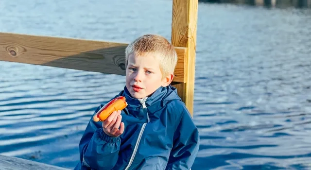 En ung pojke sitter på en brygga och äter en korv