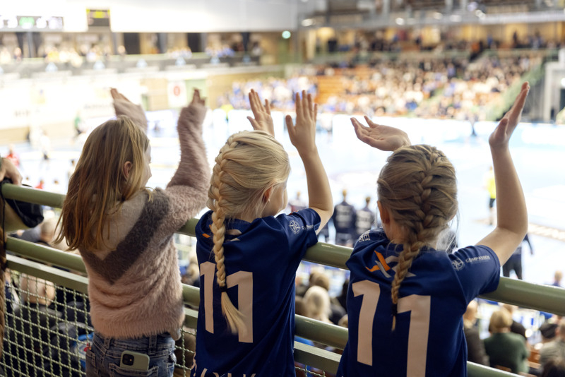 Tre flickor står och kollar på handbollsmatch.