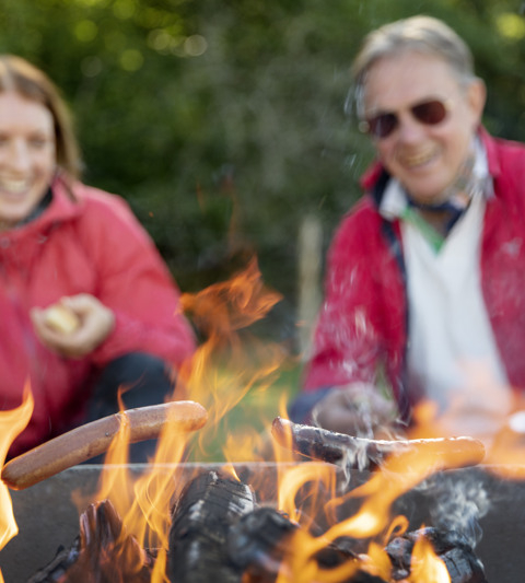 Younger woman and older man grilling sausages, with the fire in focus.
