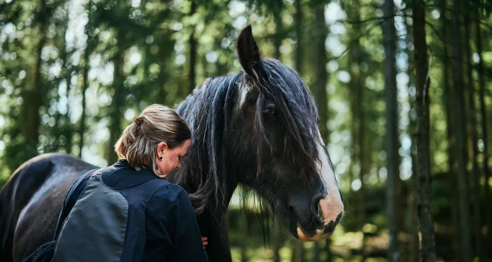 En kvinna står bredvid en häst i en skog.