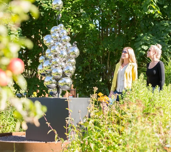 Två kvinnor som står och kollar på en skulptur med massa silvriga runda bubblor. Grönt runt om kring.