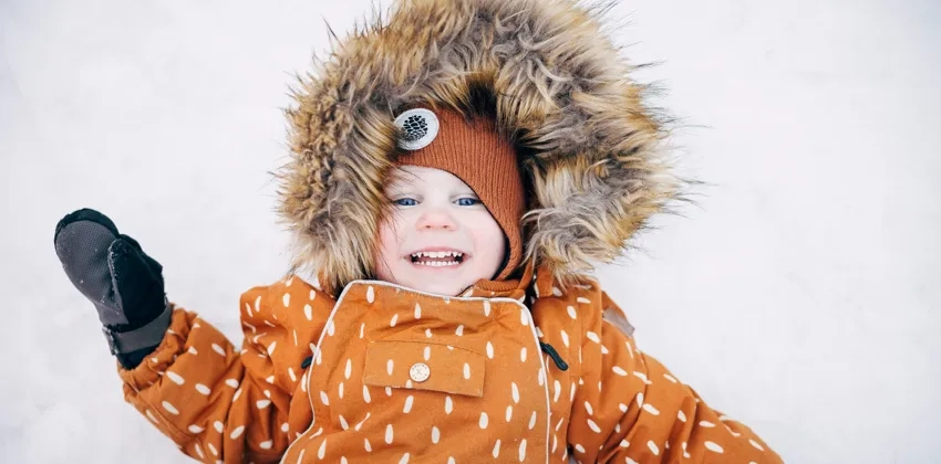 Ett barn i vinteroverall ler i snön.
