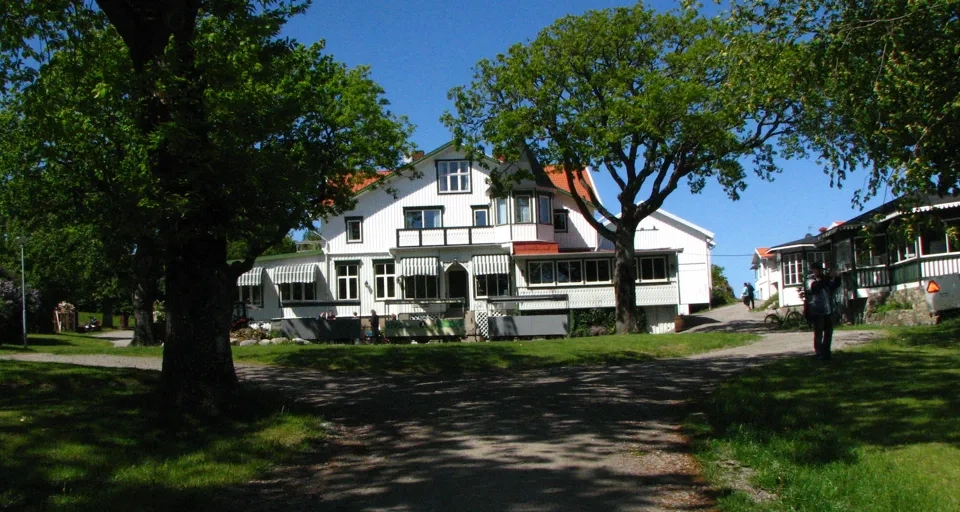 Hotel Koster Ekenäs, vit träbyggnad med grönvita markiser och högväxta ekar framför hotellet.