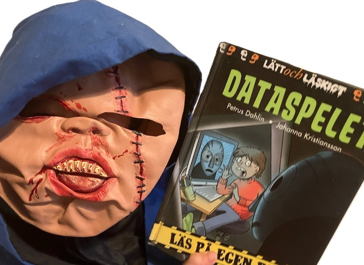 En person iförd skräckmask håller i boken Dataspelet av Petrus Dahlin