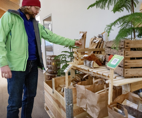 En man i grön jacka står bredvid en hög med trälådor och påsar med vedklampar.