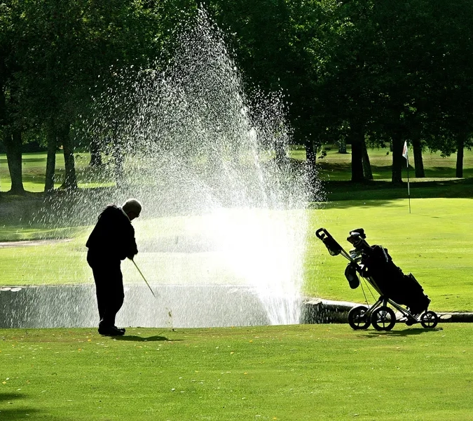 En man står på en green och golfar. I bakgrunden syns en fontän spruta och bredvid honom står hans golfvagn.