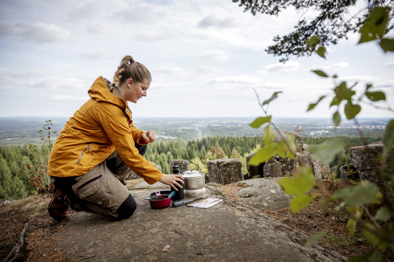 Girl cooking on a tripod stove outdoors, with a view in the background.