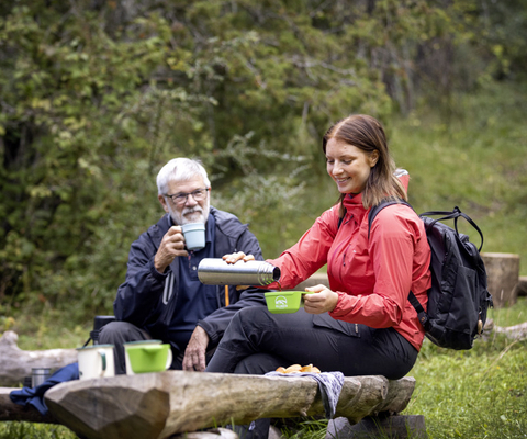 Two people sitting and having coffee on wooden benches in the forest.