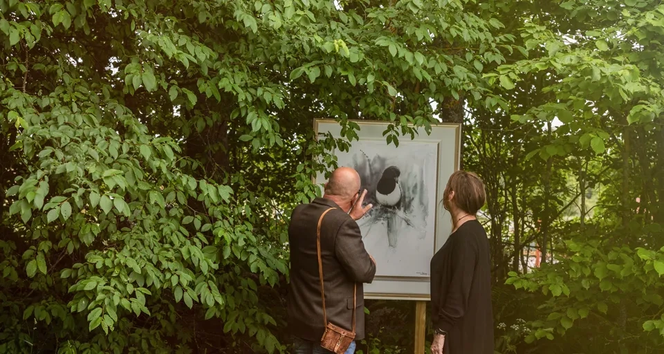 Man och kvinna som kikar på en tavla utomhus bland grönskande blad.