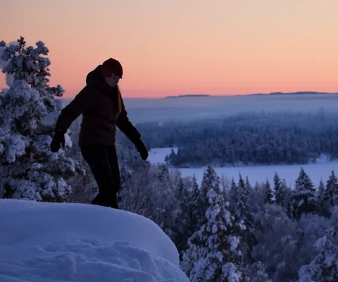 Kvinna vandrar i skymningen i snöklädd skogsmiljö med utsikt över en sjö