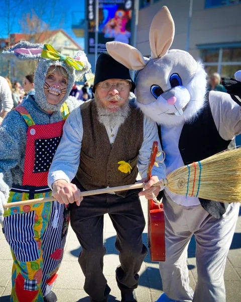 På rad står två påskharar med en påskgubbe i mellan sig. De håller en kvast framför sig. Bakom som syn ett folkvimmel.