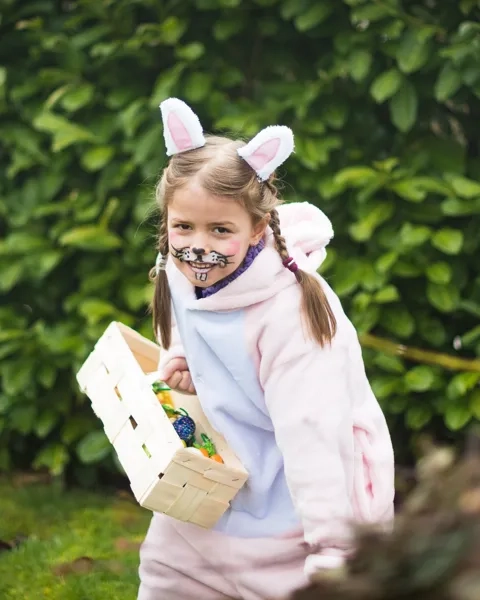 Ung flicka iklädd rosavit kanindräkt påväg mot kameran, håller i en korg med påskägg