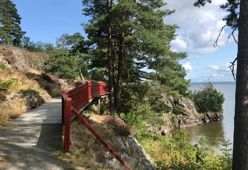 En röd träbro längs med Vänerns kust. I bakgrunden skymtas Kinnekulle. Det är en solig vårdag innan trädens knoppar brustit ut.