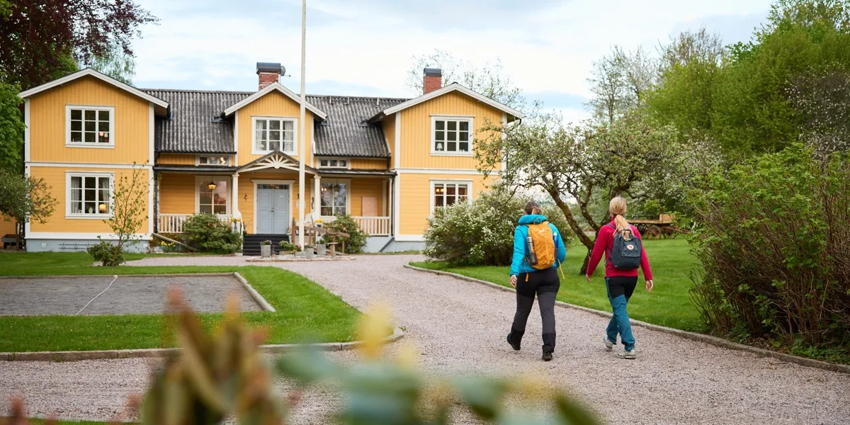 Två vandrare promenerar på uppfarten mot ett gult pensionat.