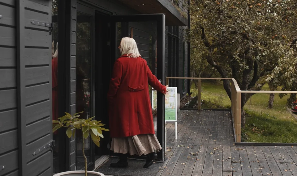 Kvinna i röd kappa går in i ett svart trähus.