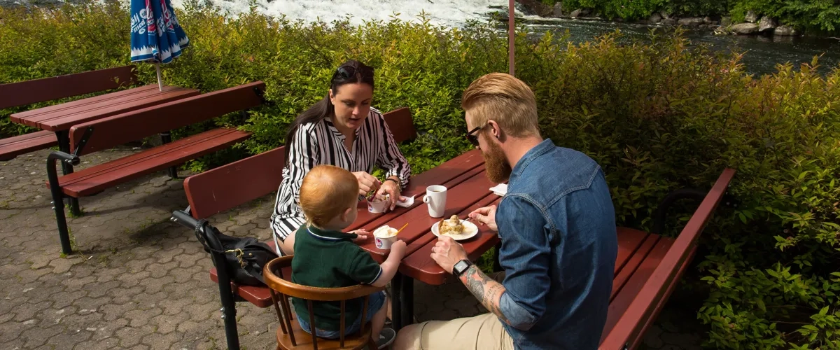 Två vuxna och ett barn sitter och fikar på terassen utmed ån Tidan på Turbinhusön i Tidaholm en sommardag.