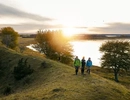 Tre personer vandrar på en ås i solnedgången. Bakom dem syns Hornborgasjöns blanka vatten.