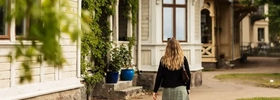 A woman walking down a dirt road in front of a house.