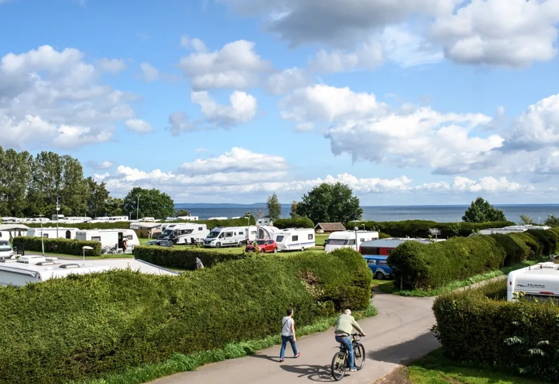 Översikt över Hjo camping, man ser två personer cykla och en massa husvagnar och husbilar som står bakom en stor häck. Vattnet skymtas längre bort.