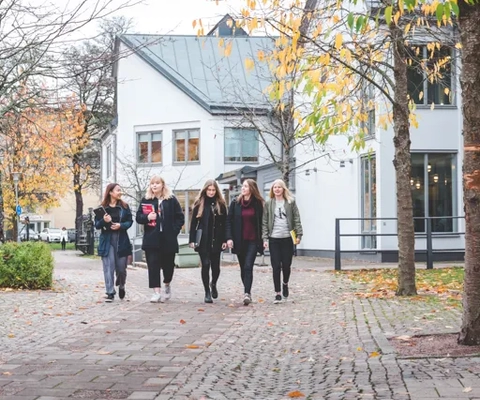 Fem kvinnliga elever med böcker i händerna promenerar från en vit byggnad med grönt tak. Runt dem syns flera träd med gula, orange och gröna löv.