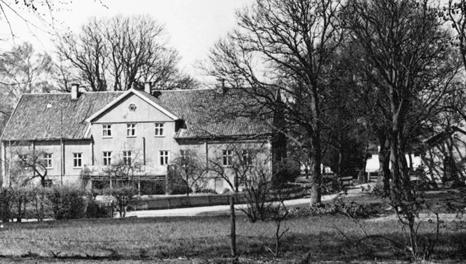 En ljus byggnad i bakgrunden på en gård. En gräsmatta med träd i förgrunden.