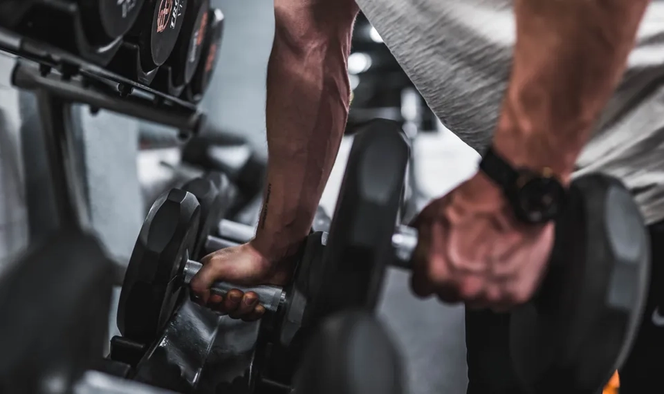 En närbild på två manliga underarmar som tar tag i två hantlar på ett gym.