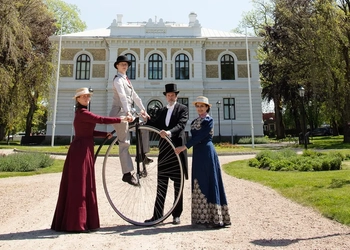 Fyra personer i 1800-tals kläder. En sitter på en höghjuling.