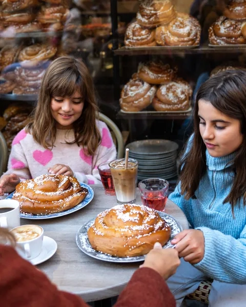 Två flickor som sitter runt ett bord, utomhus på ett kafé och dricker saft och äter kanelbullar