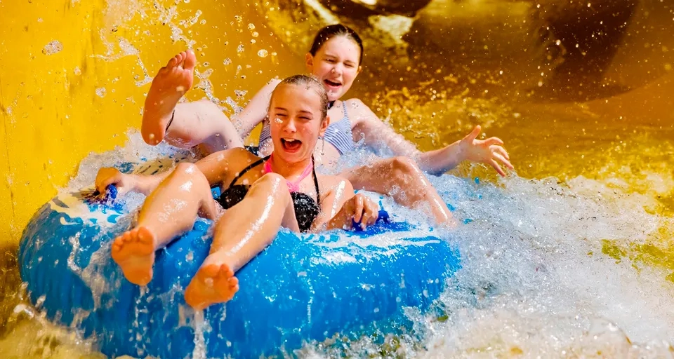 Två superglada tjejer åker på en blå badring i en gul vattenrutschkana så att vattnet stänker.
