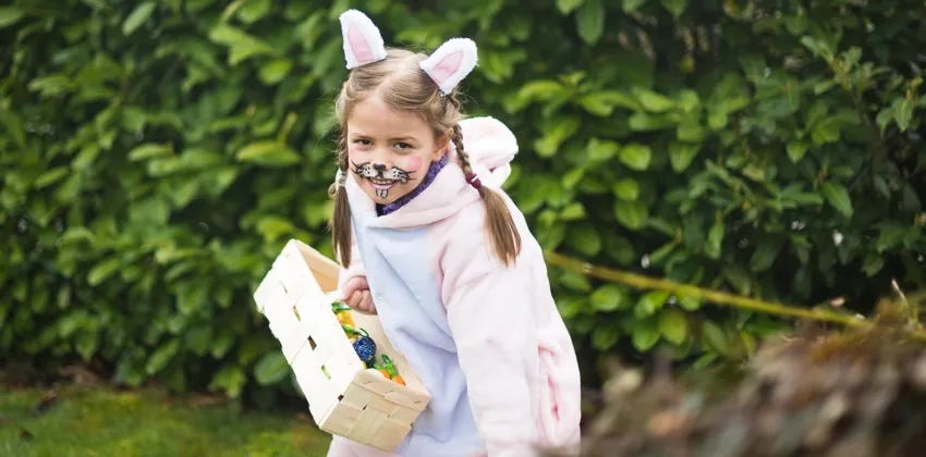 Ung flicka iklädd rosavit kanindräkt påväg mot kameran, håller i en korg med påskägg