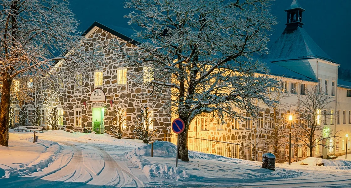 En bild på en gammal vacker fabrik med stenfasad. På tak, mark och träd ligger ett tjockt snötäcke. Himlen är midnattsblå.