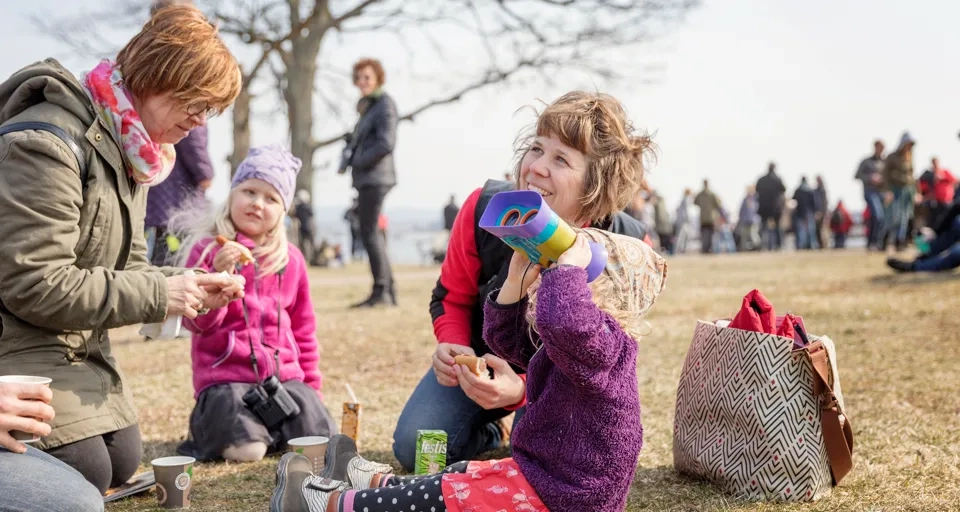 Barn och vuxna som sitter på marken och fikar. Ett av barnen kikar mot himlen med kikare.