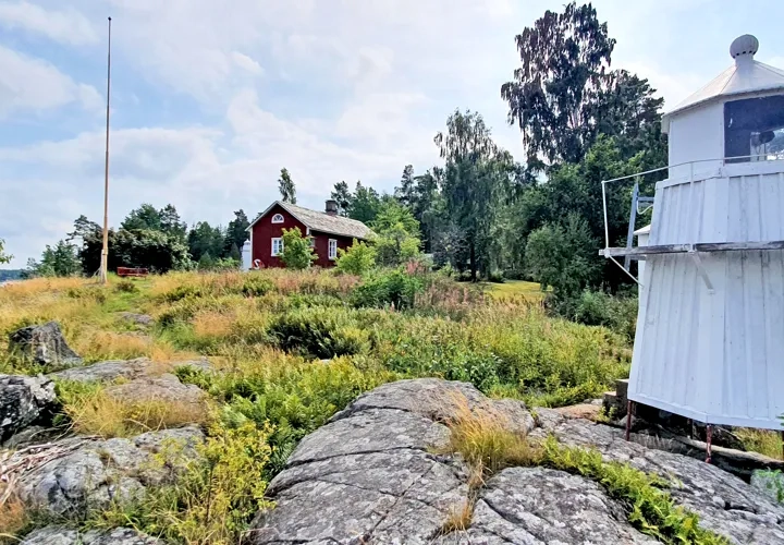 Fyrplatsen i Harnäs med rött boningshus med liten fyrlykta på fasaden, en vit fyr och Vänern i bakgrunden.