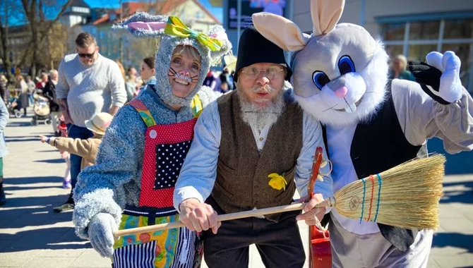 På rad står två påskharar med en påskgubbe i mellan sig. De håller en kvast framför sig. Bakom som syn ett folkvimmel.