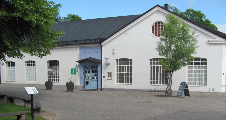 Tidaholms museums entré som är en vit stenbyggnad med en blå dörr i sommarskrud med en kullerstensgata framför.