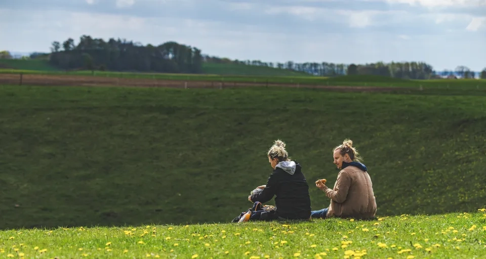 Två personer sitter på kanten till en dödisgrop täckt av grönt gräs. I förgrunden syns gula maskrosor.