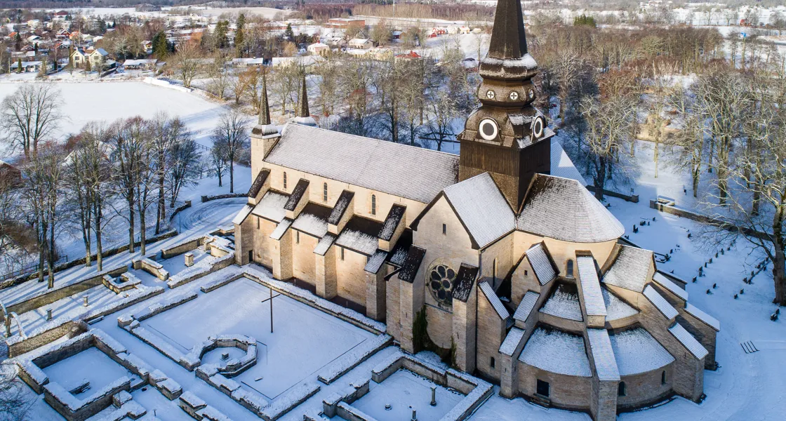 Drönarbild över Varnhems klosterkyrka med snö på taket och det omkringliggande landskapet.