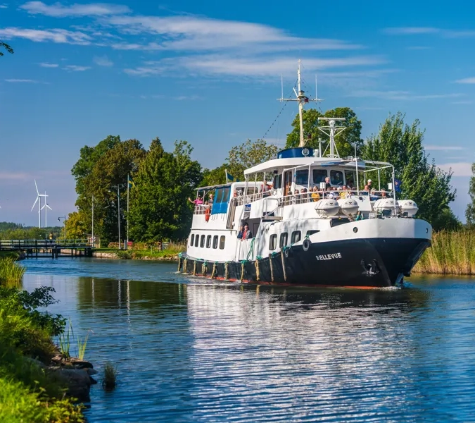 A large boat traveling down a river next to a forest.
