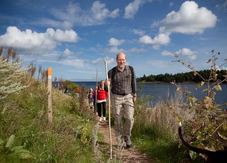 En man i förgrunden och bakom sig har hon ett led med vandrare. Stigen går längs med vattnet.