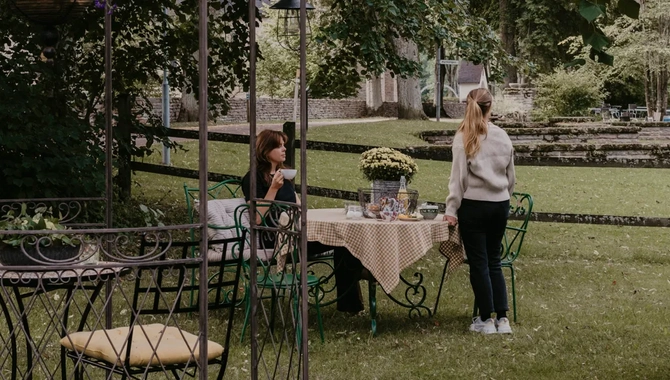 Två kvinnor sitter och står vid ett bord i en trädgård. Den ena kvinnan håller i en kaffekopp.