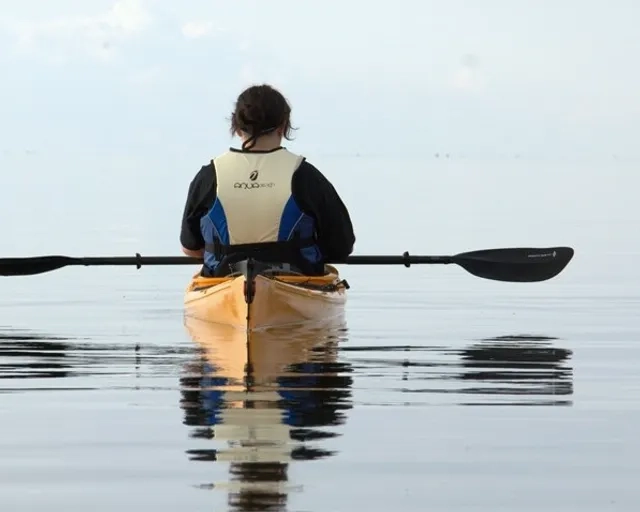 En person sitter i en kajak med ryggen mot kameran. Vattnet är stilla och går i samma gråblåa ton som himlen.