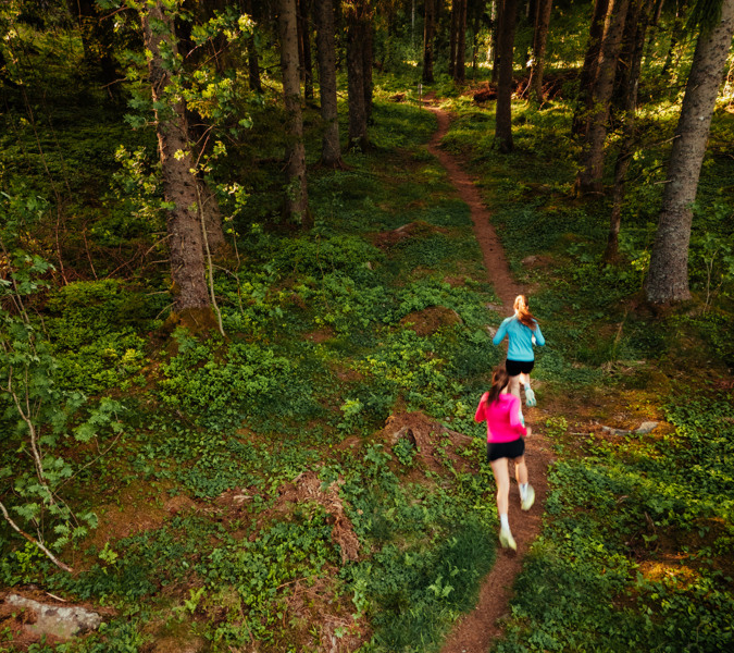Two women running through a forest on a trail.