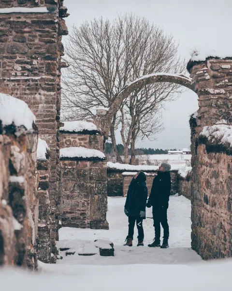 Snöigt landskap. Två personer står och kikar uppåt på en valvbåge i ruinerna av klosterkyrkan i Gudhem.