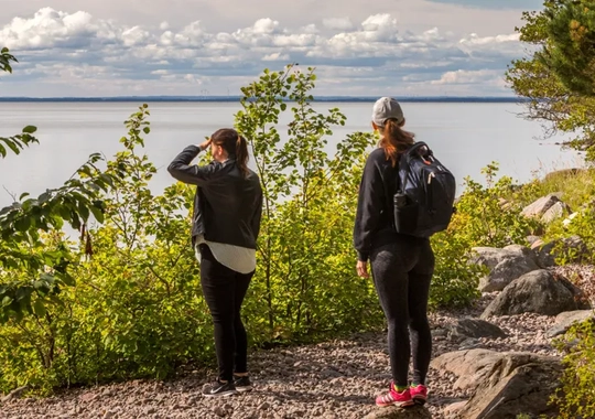 Två personer står och tittar ut mot vatten (sjön Vänern). Marken de står på består av grus och sten. Runt omkring dem syns gröna buskar och träd. Den ena personen bär på en ryggsäck och är klädd i ett par rosa joggingskor och en grå keps. Den andra personen bär ett par svarta joggingskor och en skinnjacka.