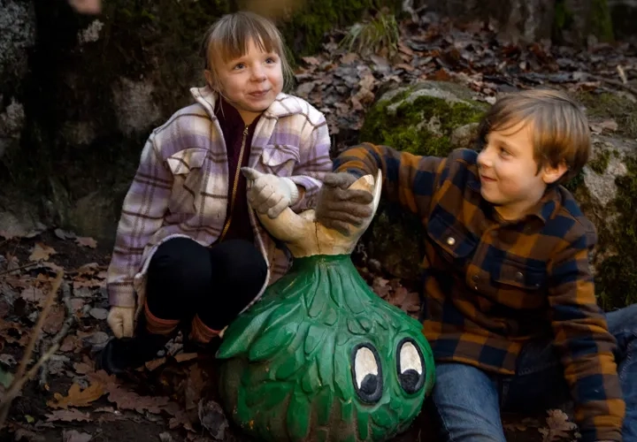 Två barn i rutiga ytterkläder sitter på ett berg med en grön figur framför sig