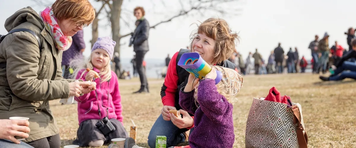 Barn och vuxna som sitter på marken och fikar. Ett av barnen kikar mot himlen med kikare.