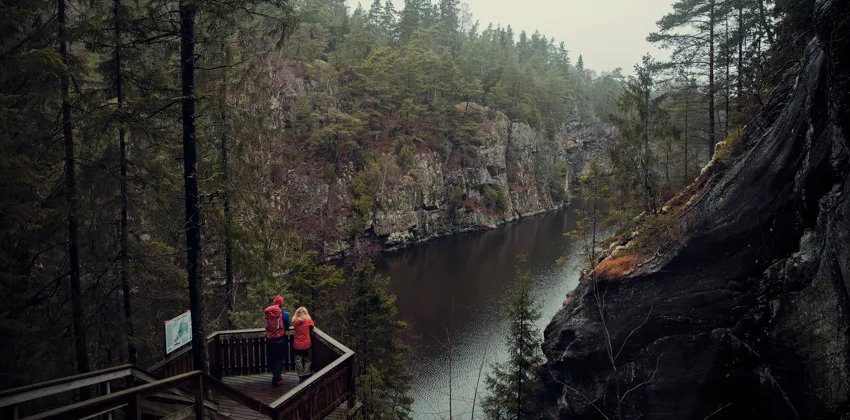 Två vandrare står på en plattform i trä och tittar ut över ett smalt sund med branta bergsväggar på sidorna