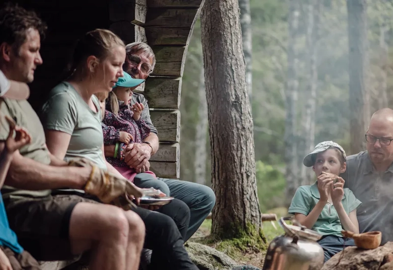 Vuxna och barn som sitter i och vid ett vindskydd och fikar.